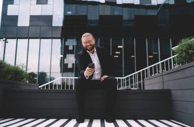 Gözlüklü, resmi takım elbiseli, kameraya bakıp gülümseyen, cep telefonundaki uygulamayı kullanan ve uzaktan çalışan mutlu sakallı bir erkek.