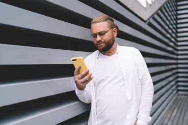 Gözlüklü, cep telefonu ekranına bakan ve gün ışığında merdiven inşaatının yanında durup mesaj okuyan sakallı genç bir erkek.