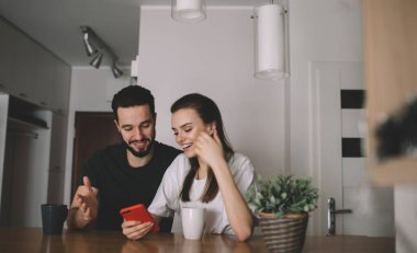 Moda kıyafetleri içinde, cep telefonuyla iletişim kuran ve rahat bir mutfakta el kol hareketleri yapan mutlu bir genç adam ve kadın.