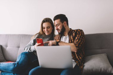 Oturma odasındaki dizüstü bilgisayarla rahat bir kanepede dinlenirken gülümseyerek ve akıllı telefonu paylaşarak mutlu bir çift.