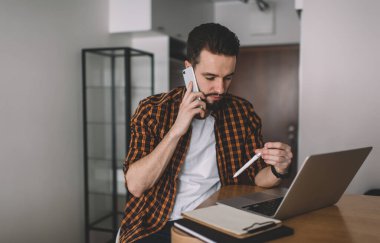 Rahat gömlekli ciddi bir erkek girişimci dizüstü bilgisayarla masada oturuyor ve iş programını cep telefonuyla tartışıyor.