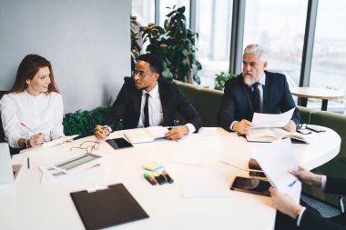 Bir grup çok ırklı iş arkadaşı resmi giysiler içinde masa başında kağıt ve belgelerle otururken aynı zamanda modern konferans salonunda müdürün masasında birlikte çalışıyorlar.