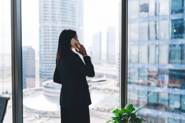 Cam duvarın yanında duran ve iş planlarını iş yerinde cep telefonuyla tartışan resmi kıyafetli kadın girişimcinin yan görüntüsü