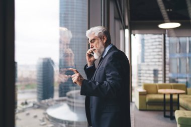 Şık takım elbiseli yaşlı bir girişimcinin modern şehrin penceresinin yanında dururken cep telefonuyla konuşmasının yan görüntüsü.