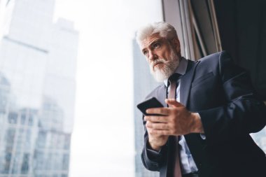 İş yerinde büyük bir pencerenin yanında dikilirken elinde cep telefonu tutan gri saçlı, ciddi bir erkek.
