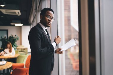 Resmi takım elbiseli ciddi bir Afro-Amerikan erkek girişimcinin yan görünüşü. Pencere kenarında çalışıyor. Ofis evrakları ve kameraya bakıyor.