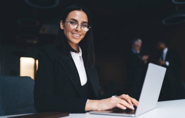 Siyah takım elbiseli ve gözlüklü pozitif kadın ofis çalışanı, karanlık arka planda iş arkadaşlarıyla masada otururken netbook 'ta yazı yazıyor.