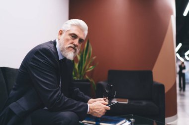 Siyah koltukta oturmuş, iş yerinde kameraya bakarken internette sörf yapan yaşlı, sakallı bir patron.