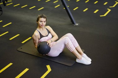 Pembe aktif giysili genç atletik kadının tüm vücudu yüksek açıyla antrenman sırasında tıp topu ile egzersiz yaparken mindere oturuyor.
