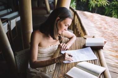 Yüksek konsantrasyonlu kadın girişimci bilgileri kontrol ederken ve notlar alırken, tropik tatil beldesindeki kafe projesiyle ilgili kağıtlarla birlikte çalışıyor.