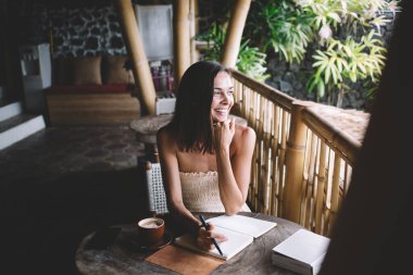 Neşeli, genç bir bayan, ahşap masada bir fincan kahve defteriyle çenesine dokunarak oturuyor ve kafeteryada elinde kalemle başka yere bakıyor.