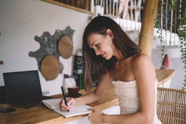 Sakin, yaz kıyafetleriyle genç bir bayan dizüstü bilgisayarla ahşap masaya otururken ve kafeden projeye uzaktan yakından bakarken deftere fikirler yazıyor.