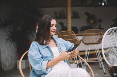 Yandan bakan mutlu genç bayan ekranda cep telefonuna bakıyor ve kahve fincanı ile sandalyede oturuyor ve açık kafede güneş ışığında dinleniyor.