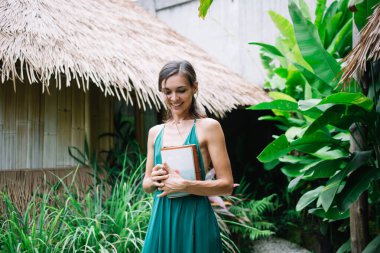 Zarif bir yaz elbisesi içinde, elinde defteriyle egzotik bitkilerin yanında dururken gülümseyen ve aşağıya bakan genç bir kadın.