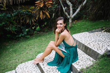 Yüksek açılı, çıplak ayaklı, son moda elbiseli, taş merdivenlerde oturmuş kameraya bakan ve gülümseyen genç bir kadın.