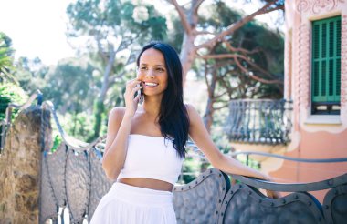 Beyaz yaz kıyafetleri içinde neşeli etnik kadın gülümseyen ve cep telefonuyla konuşan, manzaraya hayran olan ve tropik tatil beldesinde dinlenen.