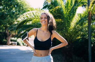 Güneşli bir günde ellerin palmiye ağaçlarının yanında dikilirken ve tatilin tadını çıkarırken akıllı telefonlu, gülümseyen ve yüzünü çeviren genç, pozitif İspanyol kadın.