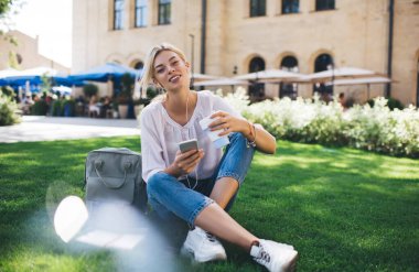 Kulaklıklı güzel beyaz kadın portresi. Yaz parkında ses podcast 'i dinlerken kameraya bakıyor. Milenyum boyunca akıllı telefon cihazıyla müzik listesinden keyif alan kaygısız kadın blog yazarı.