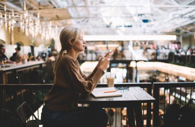 Cep telefonu kullanırken sosyal ağlarda ders kitabı sohbetleri ve mesajları olan beyaz kadın blogcuların yan görüntüsü, cep telefonu teknolojisi arama sitesinde içerik taraması yapan bin yıllık kadın.