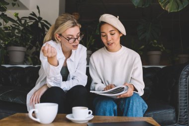 Modaya uygun bereli Asyalı bir kadın kafkasyalı bir arkadaşla görüş düzeltmesi için dergi yayınını tartışıyor moda hipster kızlar hafta sonunu kapalı mekanda konuşarak geçiriyorlar.