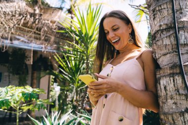 Yaz tatilinde tropikal tatil beldesinde palmiye ağacının gövdesine yaslanırken rahat giyinen, gülümseyen ve cep telefonuyla sohbet eden düşük açılı bir kadın.