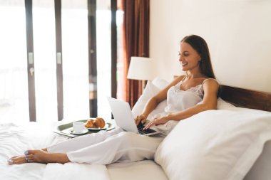 Beyaz elbiseli, yatakta oturan ve sabah kahvaltısını yaparken dizüstü bilgisayarında daktilo yazan pozitif genç bir kadın.