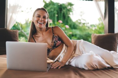 Şık kıyafetli, neşeli, genç bir bayan serbest yazar rahat bir yatakta yatarken kameraya bakıp gülümsüyor ve çardakta dizüstü bilgisayar kullanıyor.