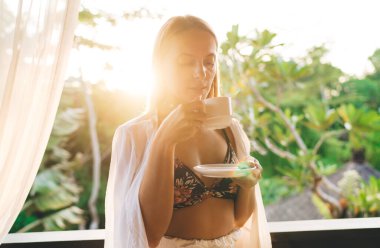 Gündüz vakti bulanık yeşil bir parka karşı ayakta duran ve elinde fincan ve fincan tabağıyla sıcak kahve içen alımlı genç bir bayan.