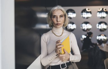 Kararlı yaşlı kadın siyah cam duvara yaslanmış ve elinde aynaları yansıtan sarı renkli kitabı tutarken kameraya bakıyor.