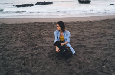 Umursayan genç bir kadın kumlu sahilde köpüklü bir köpekle otururken kayalarla köpüklü bir deniz ve bulutlu bir havada zaman geçirirken gözlerini kaçırıyor.