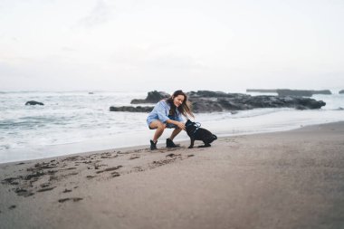 Gülümseyen genç bir bayanın kumlu sahilde dizleri bükülmüş, deniz kenarında oturup yaz günü köpek yavrusuyla oynaşırken görüntüsü.