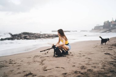 Kumlu sahilde köpüklü bir şekilde oturan genç bir kadın. Bulanık gökyüzüne ve tatil köyüne bakan uzak dalgalara bakarken köpüklü denizin yanında ellerini tutar.