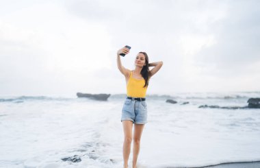 Tüm vücudu cep telefonuyla selfie çeken mutlu genç bir kadın. Ekrana bakıp saçlarına dokunurken deniz suyuna yakın duruyor ve yaz tatilinin tadını çıkarıyor.