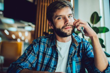 Kareli gömlekli dalgın sakallı bir erkek, yeşil saksı bitkisinin yanında otururken kameraya bakıyor ve gündüzleri bulanık kafede cep telefonu kullanıyor.