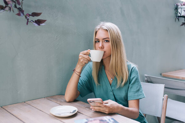 Portrait of Caucasian woman blogger with modern smartphone gadget looking at camera while drinking hot caffeine beverage in street coffee shop, millennial hipster girl with cellphone technology