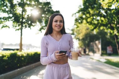 Yeşil ağaçların arasında ara sokakta duran gülümseyen genç bir kadın ve güneşli bir günde ferahlatıcı buzlu latte içerken cep telefonuyla internette gezinen.