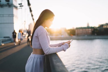 Sıska, mor kıyafetli genç bir bayanın nehir kenarındaki setin üzerinde durup gün batımında cep telefonuyla sosyal medyaya bakması.