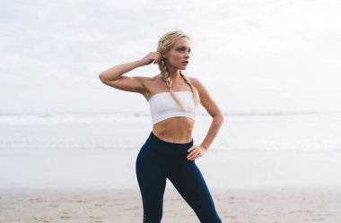 Spor kıyafetleri içindeki düşünceli fit kız deniz kıyısı sahilinde yoğun antrenman yapmayı düşünüyor, pilates yaptığı için dalgın, beyaz kadın sağlıklı yaşam tarzını zindelik için koruyor.
