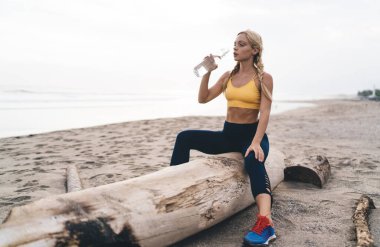 Aktif giyimli sakin bir beyaz kadın deniz kıyısındaki plajda spor molası sırasında ferahlatıcı bir şekilde içme suyu içiyor. Genç ve formda bir kız sporcu kardiyo çalışması için ormanlık alanda dinleniyor.