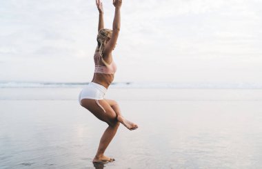 Spor kıyafetli zayıf bir kadının sabah sahilde vücut canlılığı ve esnekliği eğitimi için boş vaktinin keyfini çıkarması. Yoga yaparken kaslarını esneten sarışın kız.