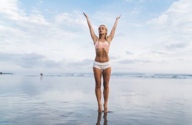 Kaldırılmış elleri olan esnek bir kadın deniz kıyısında yoga eğitimi alıyor. Canlılık sağlığına ulaşmak için gündüz vakti harcıyor. Güzel bir kadın deniz kıyısındaki plajda bütünsel iyileşme sürecinde dinleniyor.