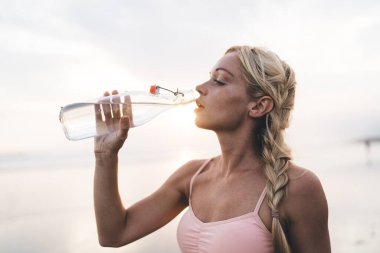 Slim Kafkas bayan atlet 20 yaşında, dinleniyor ve şişeden gelen suyla ferahlatıcı, çekici, vücudu güzel genç bir kadın deniz kıyısındaki çalışma günü boyunca soğuk bir içecek içiyor.