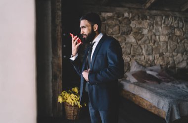 Zarif takım elbiseli ciddi bir adamın modern yatak odasında dikilip gözlerini kaçırırken akıllı telefonuyla sohbet edişinin yan görüntüsü