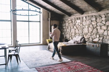 Geniş oturma odasında halıda yürüyen ve oteldeki boş vakitlerin keyfini çıkarırken cep telefonu ekranına bakan genç adamın yan görüntüsü.