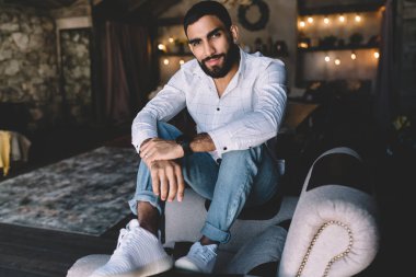 Gülümseyen sakallı erkek koltukta oturuyor ve boş zamanlarında rahat bir odada dinlenirken kameraya bakıyor.