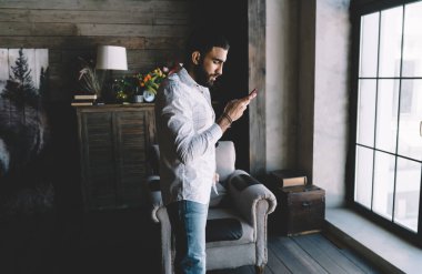 Süslü kıyafetler giyen sakallı etnik erkeğin yan görünüşü. Süslü evin penceresinin yanında durup cep telefonu kullanırken internette mesajlaşması.