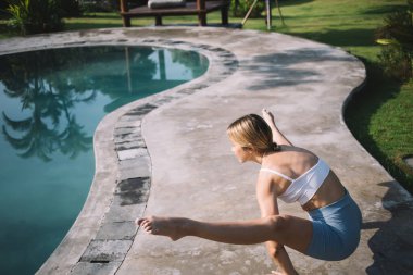 Yüksek açıda çıplak ayaklı, aktif giyimli bir kadın, güneşli bir günde havuz kenarında meditasyon yaparken ateş böceği yoga pozu veriyor.