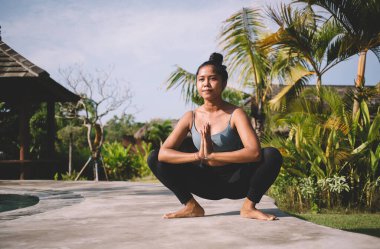 Egzotik ağaçların yanında havuz başında yoga yaparken spor kıyafetleriyle Tanrıça Asana egzersizi yapan konsantre genç etnik ayaklı bir kadın.