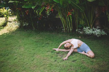 Yüksek açılı, çıplak ayaklı, spor giyimli genç bir kadın. Kapalı kapı pozu veriyor. Çimenlerde yoga yaparken gözleri kapalı.