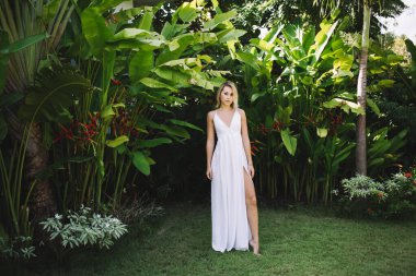 Uzun kolsuz beyaz elbise giyen, tropikal bahçede yeşil çimlerin üzerinde duran zayıf bir kadın mankeni.